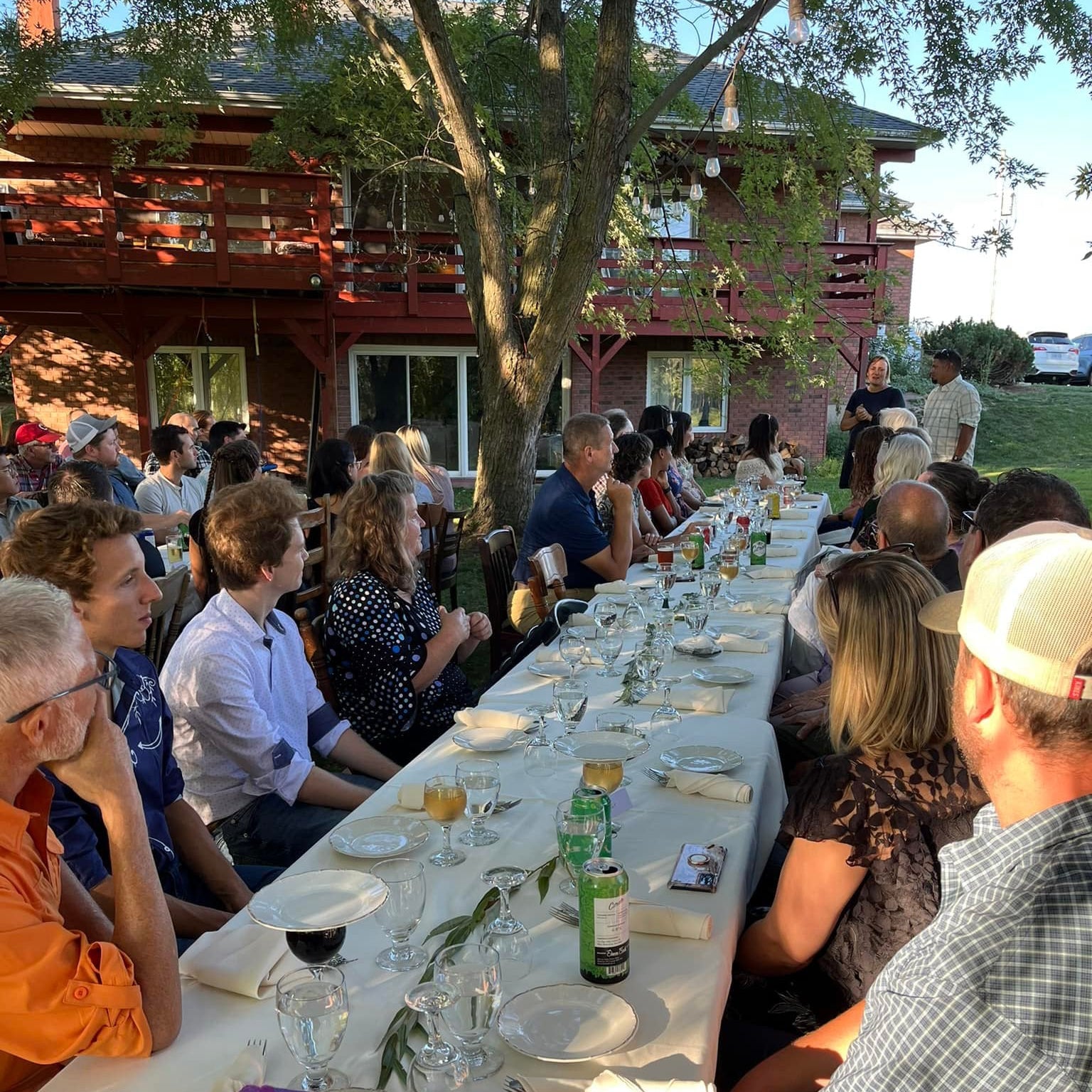 Outdoor gathering with a long table set for dinner, people seated around, and a rustic building in the background.