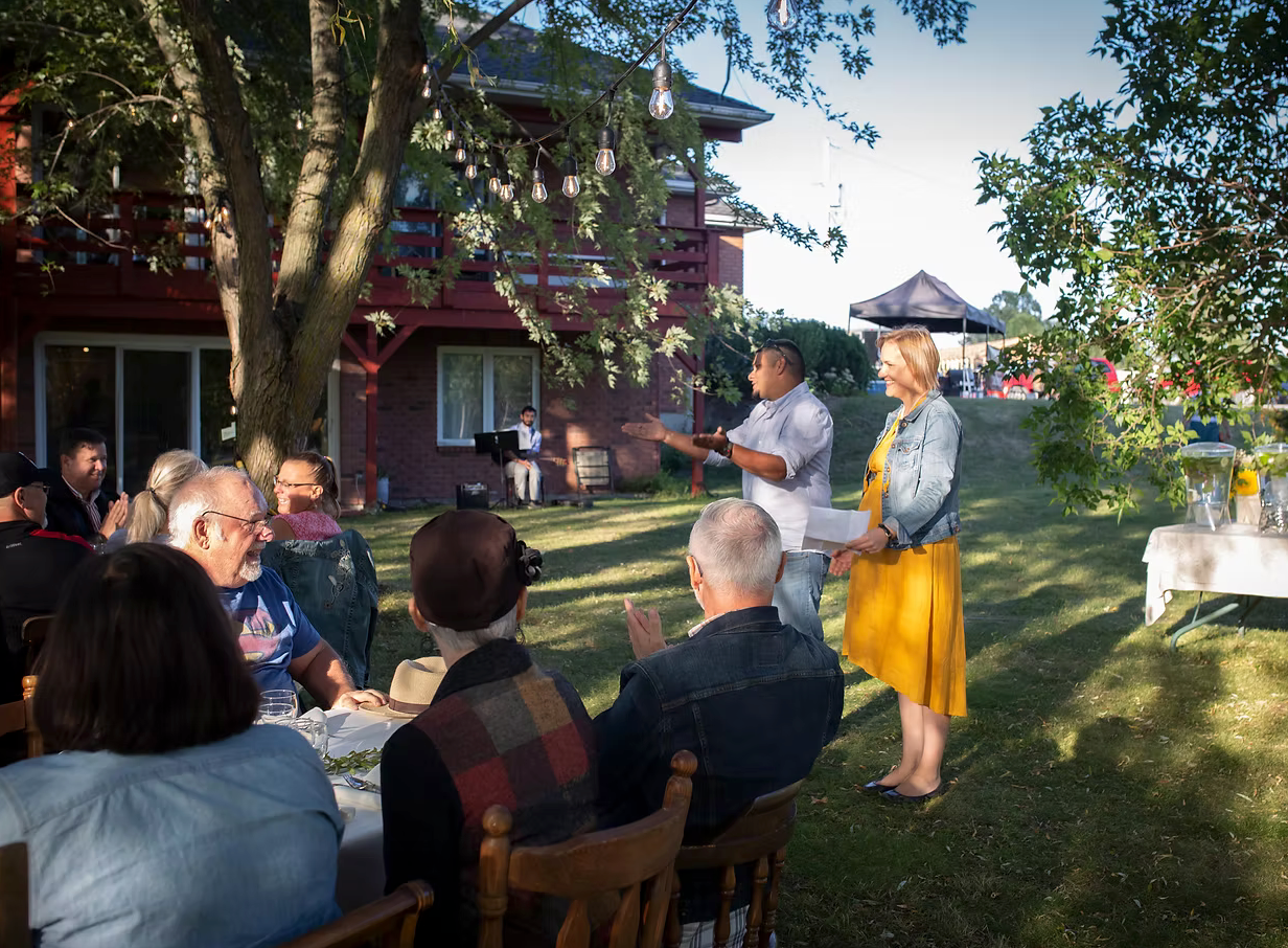 Group of people gathered outdoors for a meal, with a man in a white shirt and yellow skirt standing and speaking.