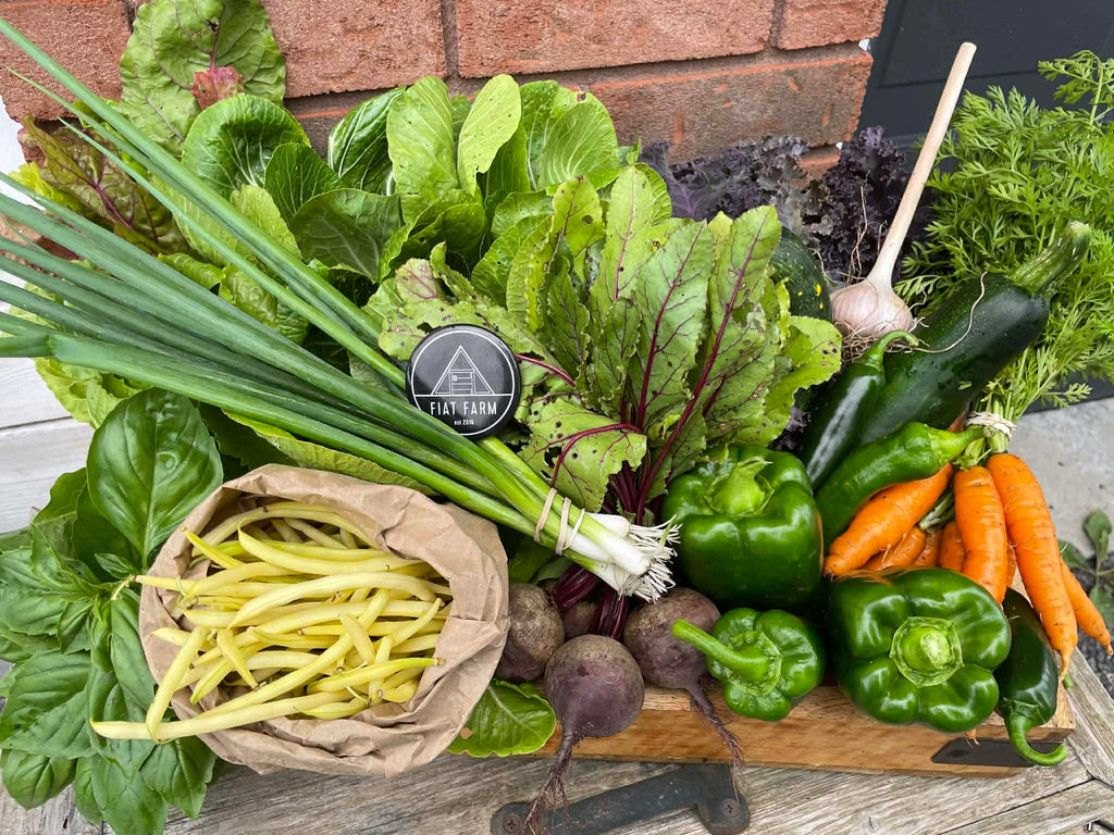 Assorted fresh vegetables including green beans, carrots, and leafy greens on a wooden board with a brand logo in the background.