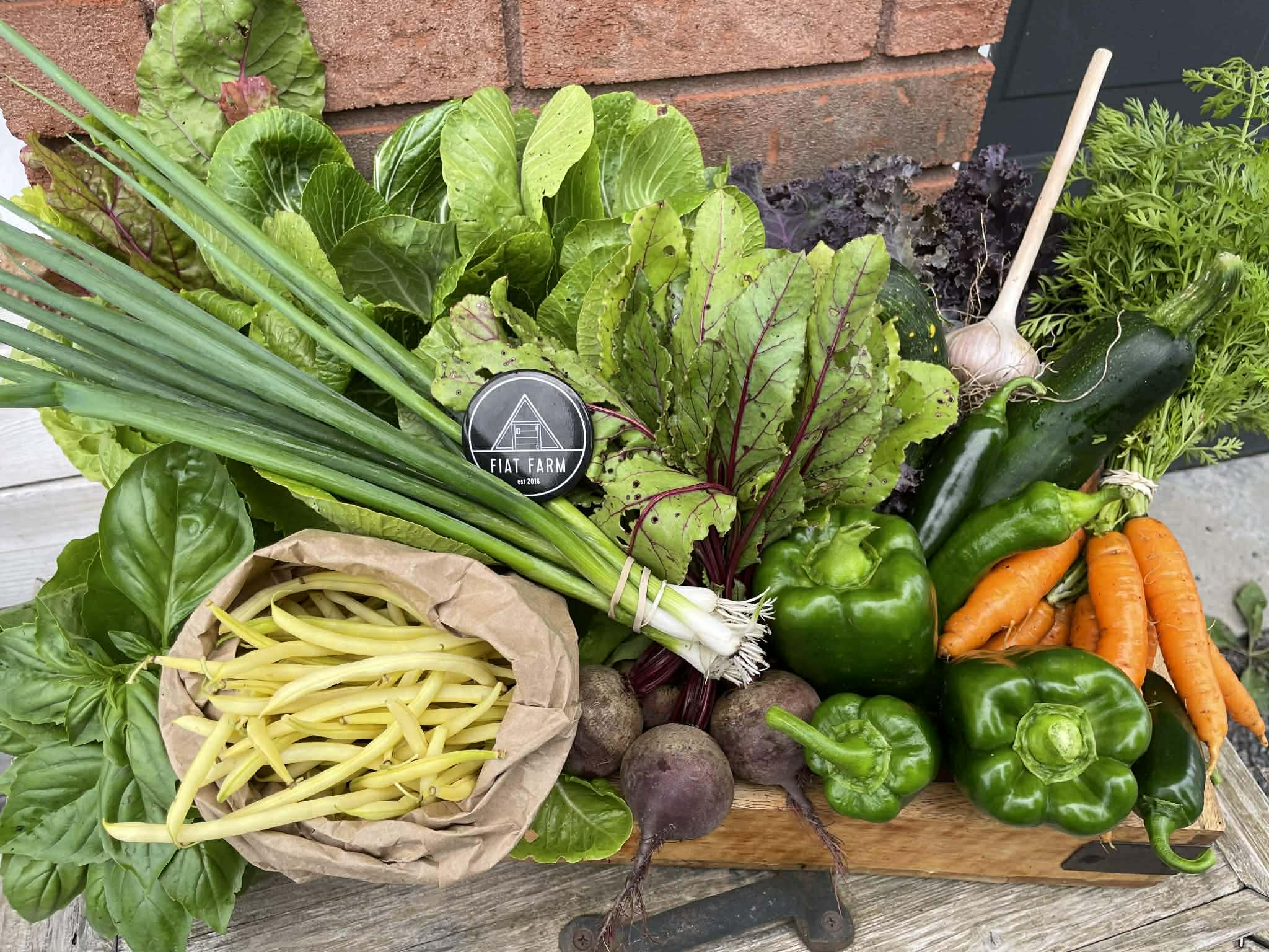 Assorted fresh vegetables including green beans, carrots, and leafy greens on a wooden board with a brand logo in the background.