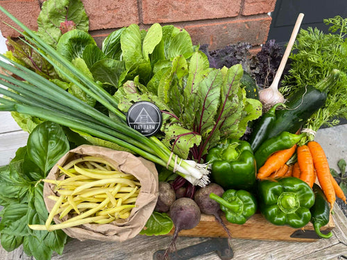 Assorted fresh vegetables including green beans, carrots, and leafy greens on a wooden board with a brand logo in the background.